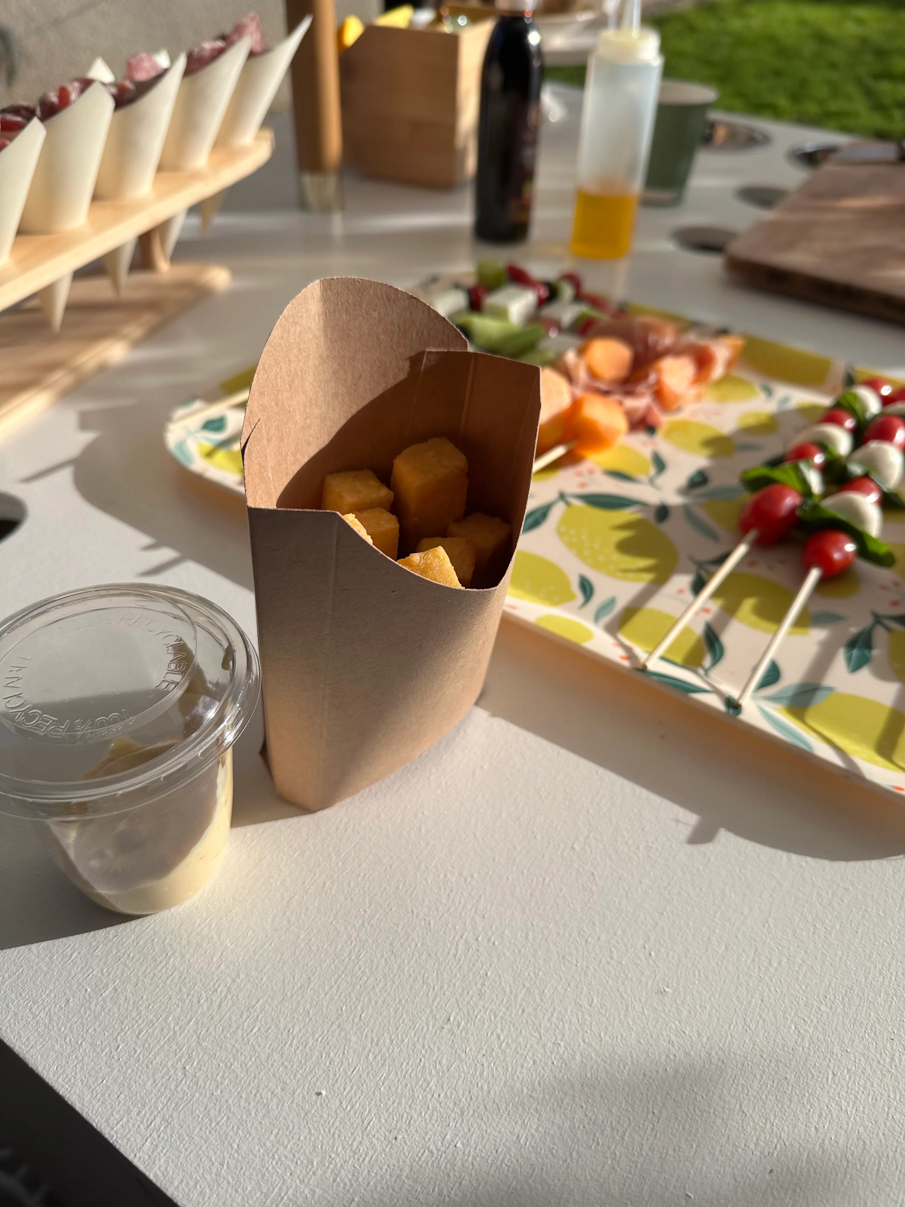 Cône kraft de polenta dorée prêt à servir, brochettes caprese (tomate, mozzarella, basilic) en arrière-plan sur nappe à motif citrons