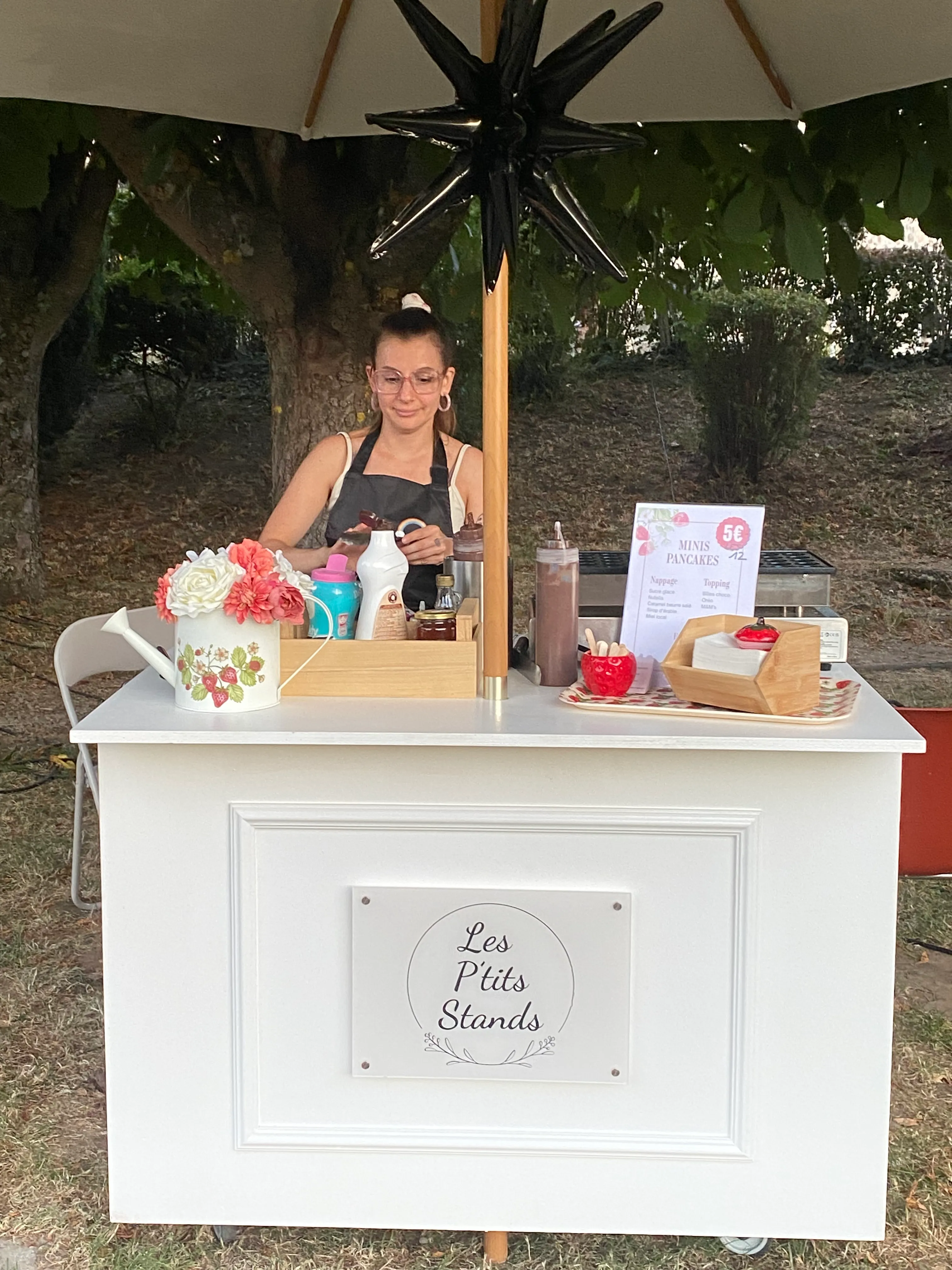 Stand sucré Les P'tits Stands installé sous son parasol en extérieur, avec le menu minis pancakes, les nappages et l'arrosoir décoratif fleuri