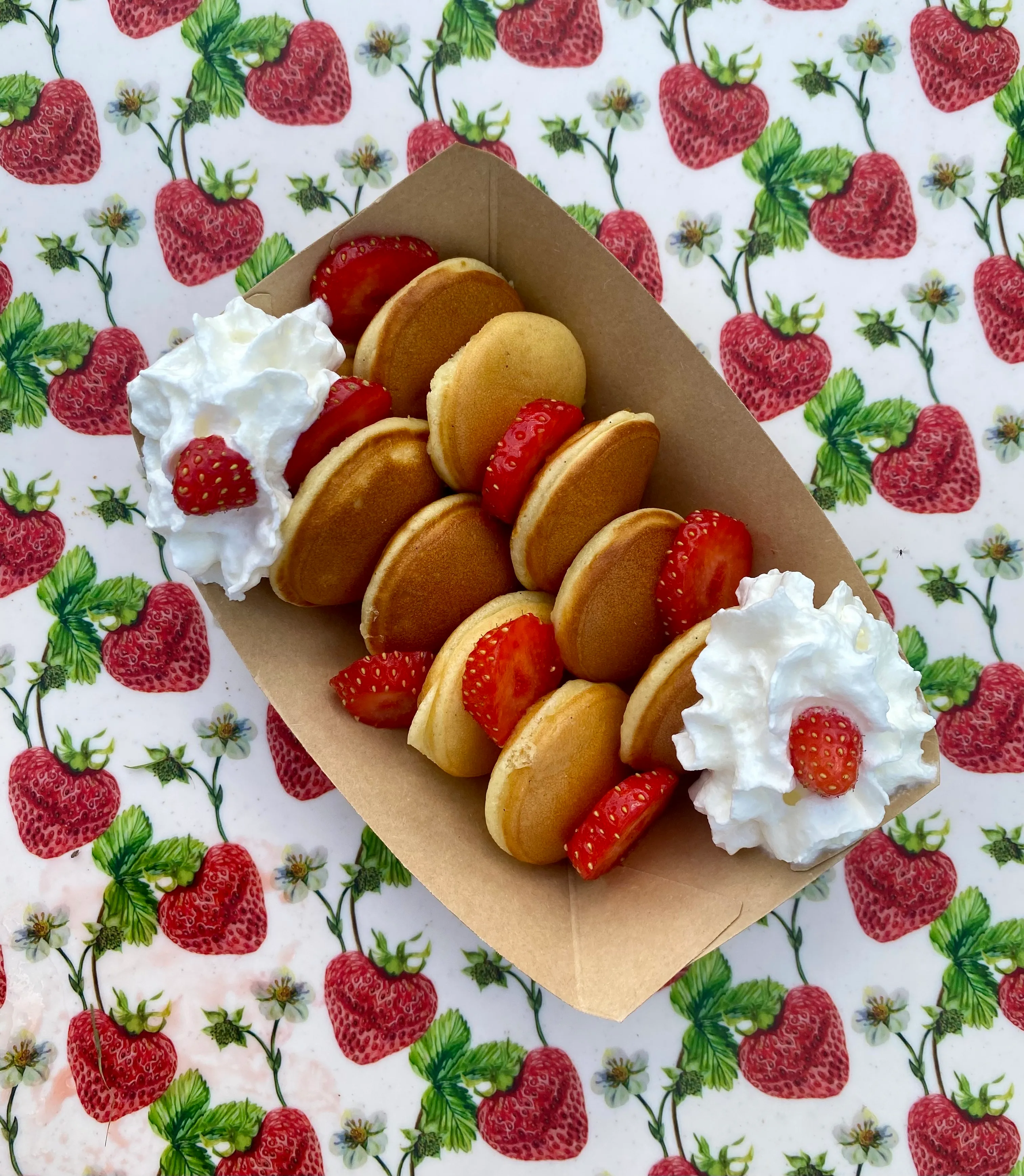 Barquette de mini pancakes garnis de fraises fraîches et de chantilly, vue plongeante sur nappe à motif fraises