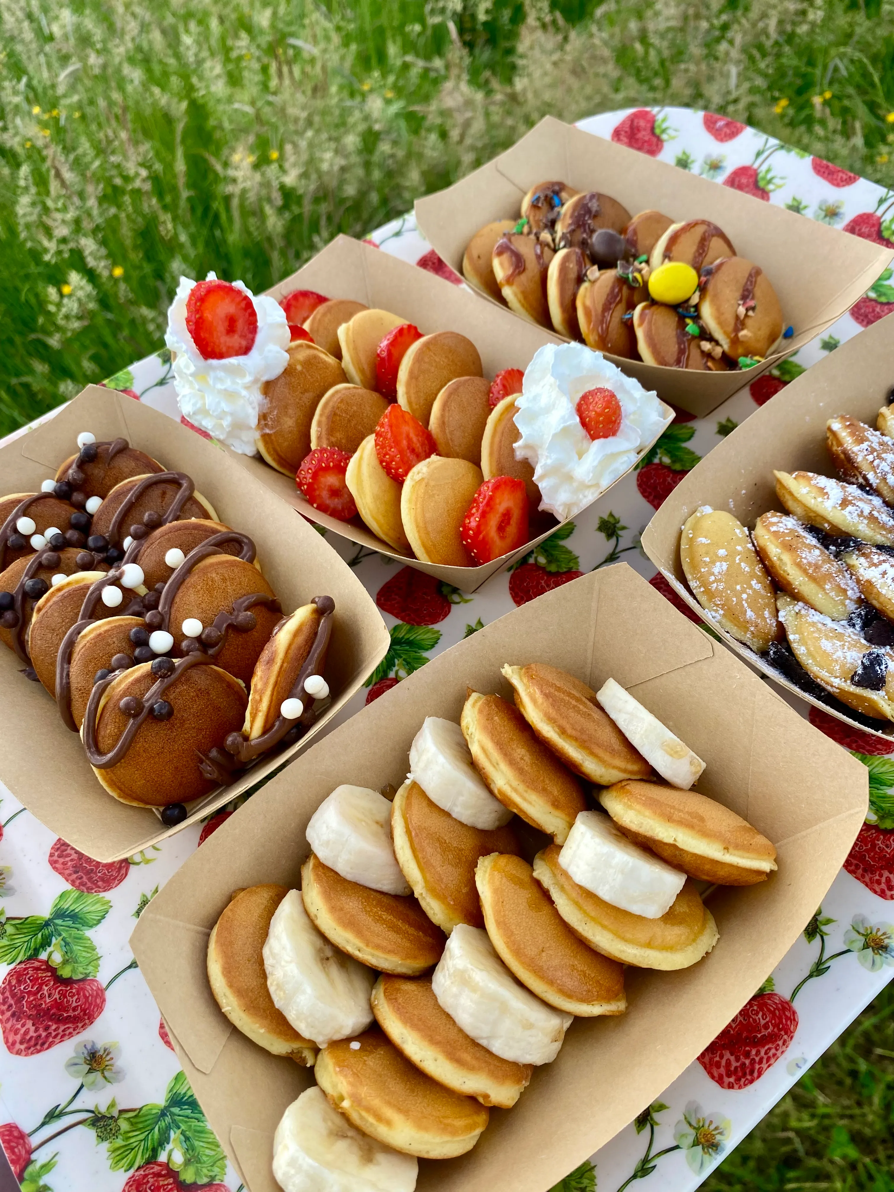 Assortiment de barquettes kraft de mini pancakes sucrés : fraises et chantilly, banane, pâte à tartiner et billes choco, présentés sur une nappe à motif fraises